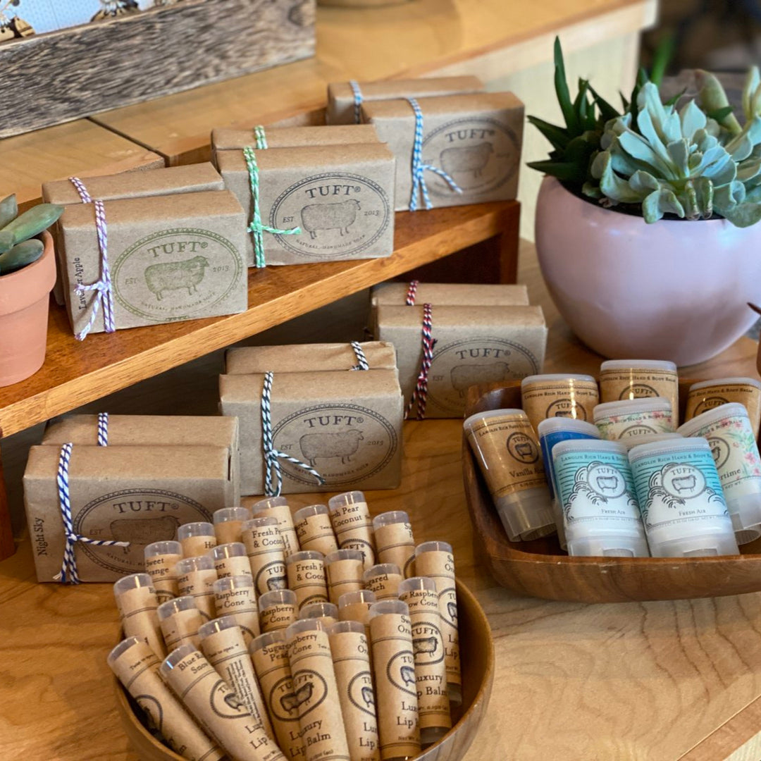 A display of various packaged soaps, with labels and tied with string, on wooden shelves alongside succulent plants.