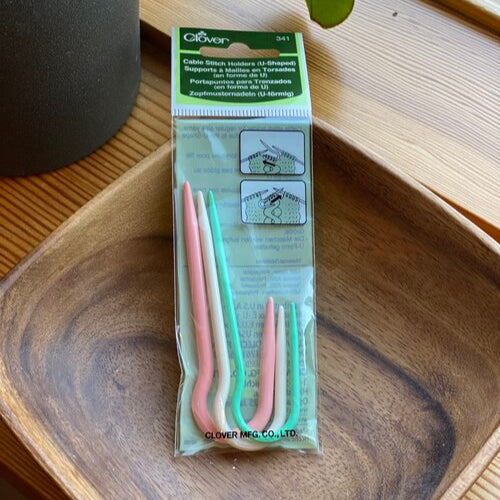 A pack of three Clover brand cable stitch holders 'J' style, displayed on a wooden tray in front of a window, with a potted plant in the background.