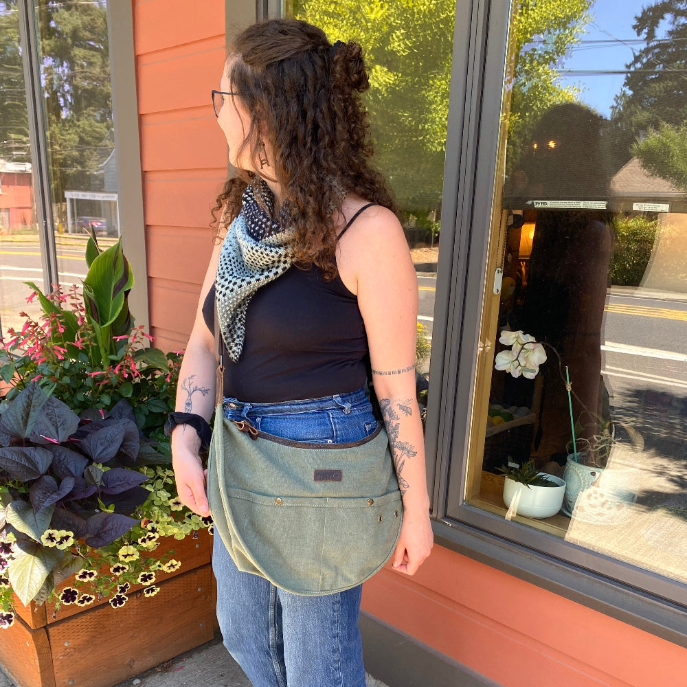 Person standing outside a building with a green bag, wearing a black top and blue jeans.