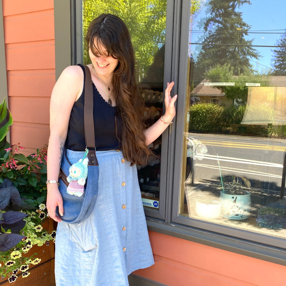 Woman standing outside a building with a window, wearing a black top, light blue skirt, and black sneakers.