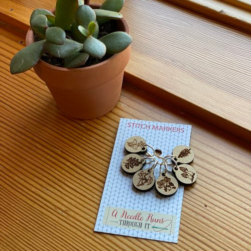 Handmade wooden stitch markers displayed on a card, with a variety of floral designs, placed next to a potted plant.