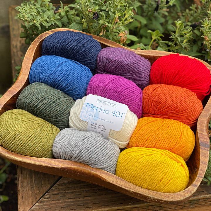 A collection of colorful yarn balls arranged in a wooden bowl, with the label 'Merino 401' visible on one of the yarns.