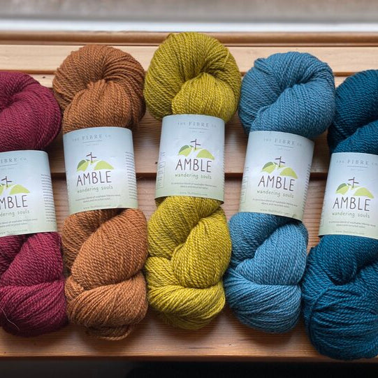 Several skeins of fingering-weight yarn in various colors are displayed, arranged in two rows with the brand label 'Amble' visible on each skein.