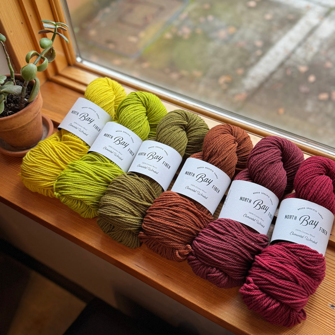 Multiple skeins of wool yarn arranged in a row, with a variety of colors including yellow, green, brown, and red, placed on a windowsill.