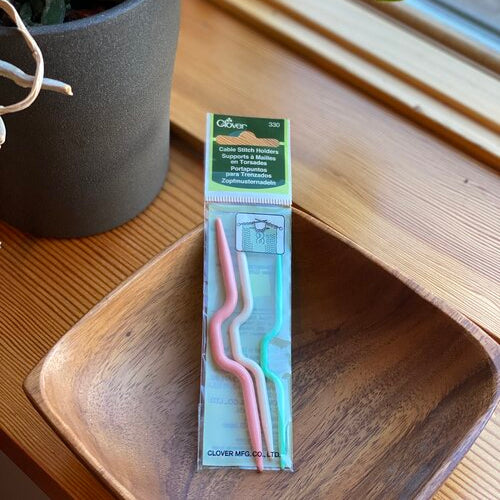 A set of cable stitch holders in various sizes placed on a wooden surface, with packaging that includes the brand name 'Clover'.