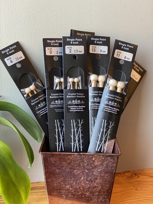 A collection of bamboo single pointed knitting needles in various sizes, displayed in a wooden box with a plant in the background.