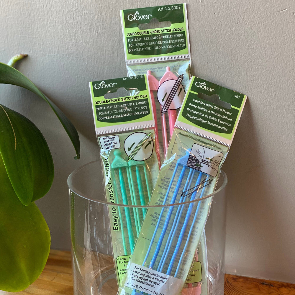 Packaging of Clover Double Ended Stitch Holders in various sizes displayed in a clear glass on a wooden surface with a plant in the background.