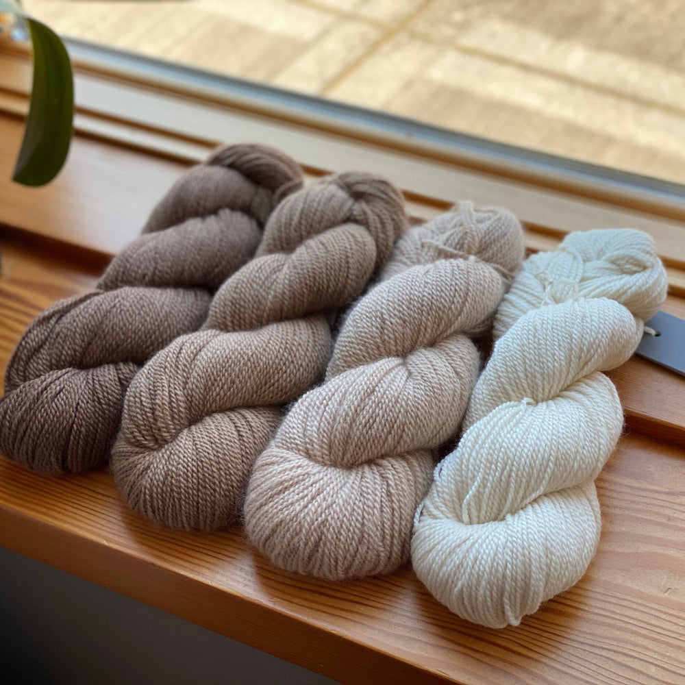 Four skeins of Tynd fingering weight yarn in beige, brown, and white colors, placed on a wooden surface.