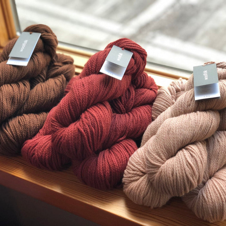 Three skeins of Woolfolk yarn in brown, red, and beige colors, placed on a windowsill.
