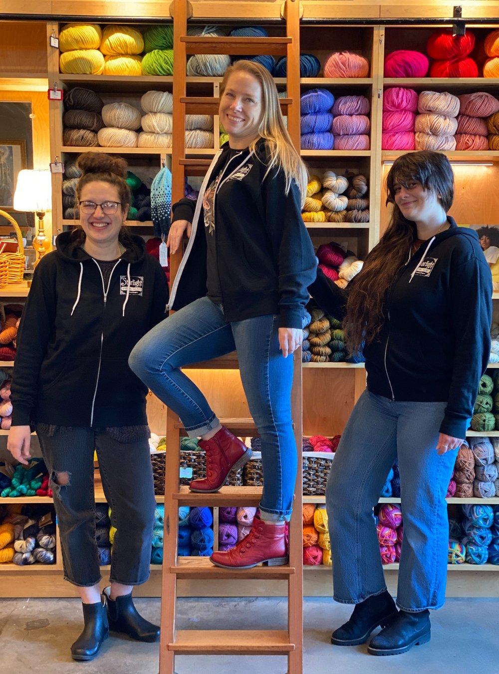 Three individuals standing in front of a wall of yarn, wearing black sweatshirts with a logo on the front.