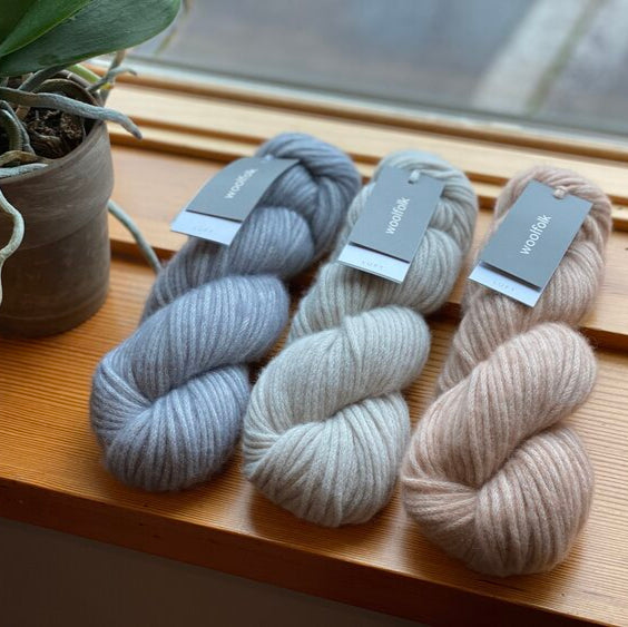 Skeins of soft squishy Luft yarn in various shades of gray and beige, displayed on a wooden surface near a window.