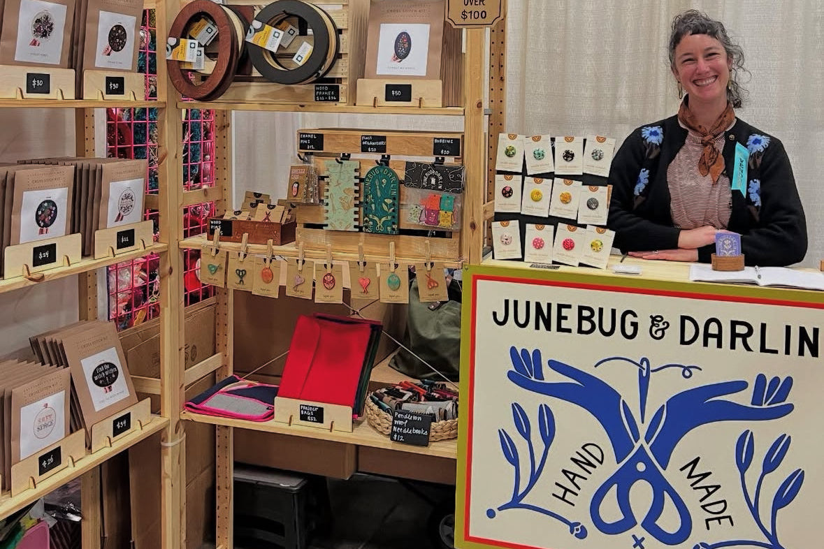 Woman standing behind a booth with 'Junebug & Darlin' branding, displaying handcrafted items.