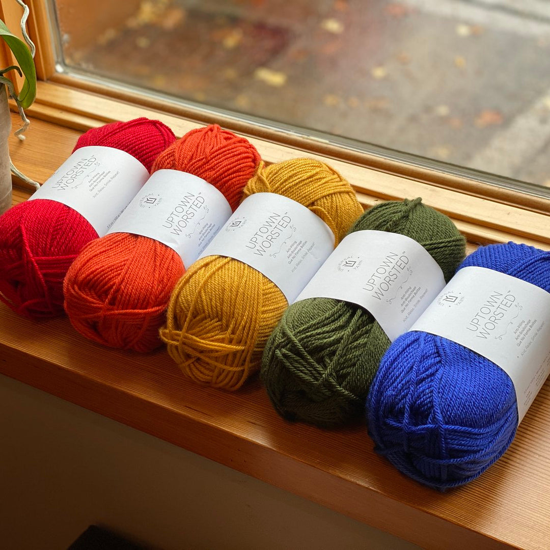 A collection of colorful yarn balls laid out on a wooden surface near a window, with a variety of colors including red, orange, yellow, green, and blue.