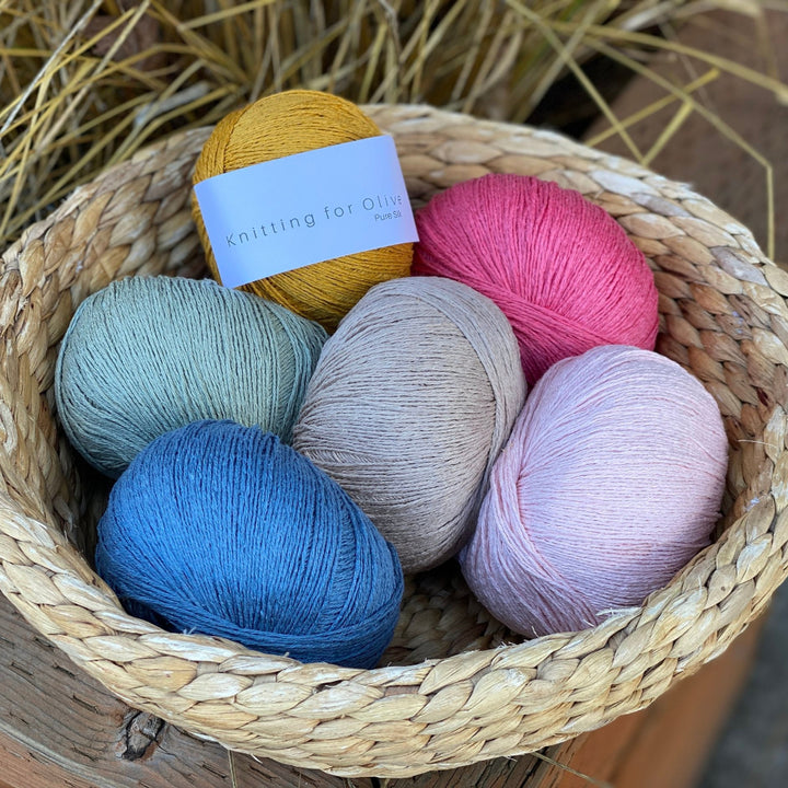 A basket containing multiple skeins of knitting for olive pure silk yarn in various colors including blue, pink, green, beige, and yellow.