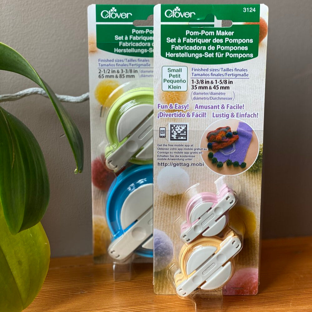 Packaging of Clover brand pom pom makers, showing two sizes of the tool with colorful yarns and finished pom-poms, placed on a wooden surface.