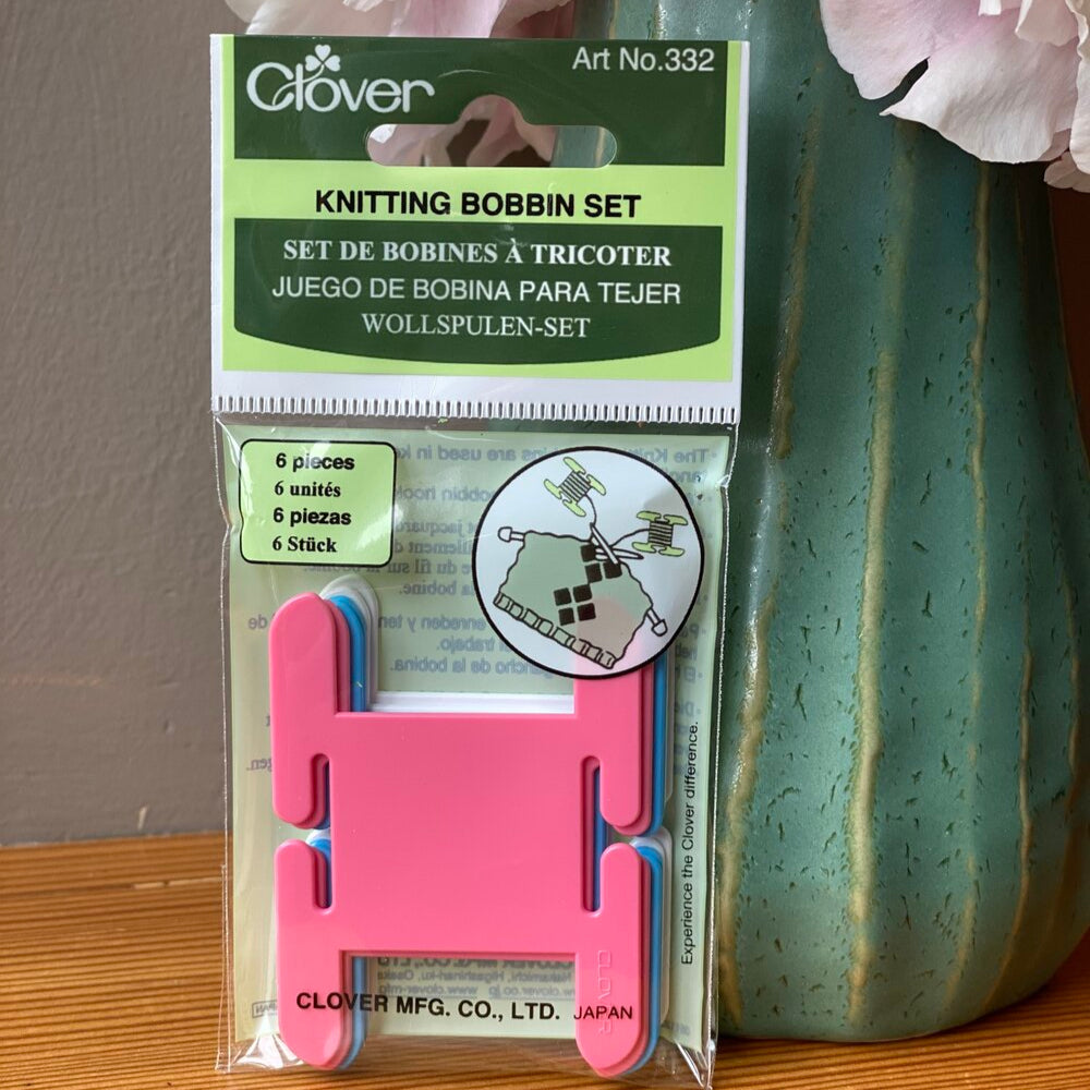 A packaged knitting bobbin set by Clover, with pink and blue bobbins visible through the clear plastic packaging, set number 332, displayed on a table with a vase of pink flowers in the background.