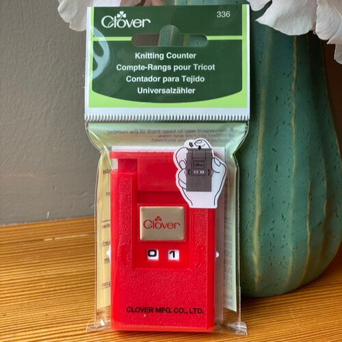 A red knitting counter by Clover, designed for keeping track of rows in knitting and crochet projects, displayed in front of a vase with flowers.