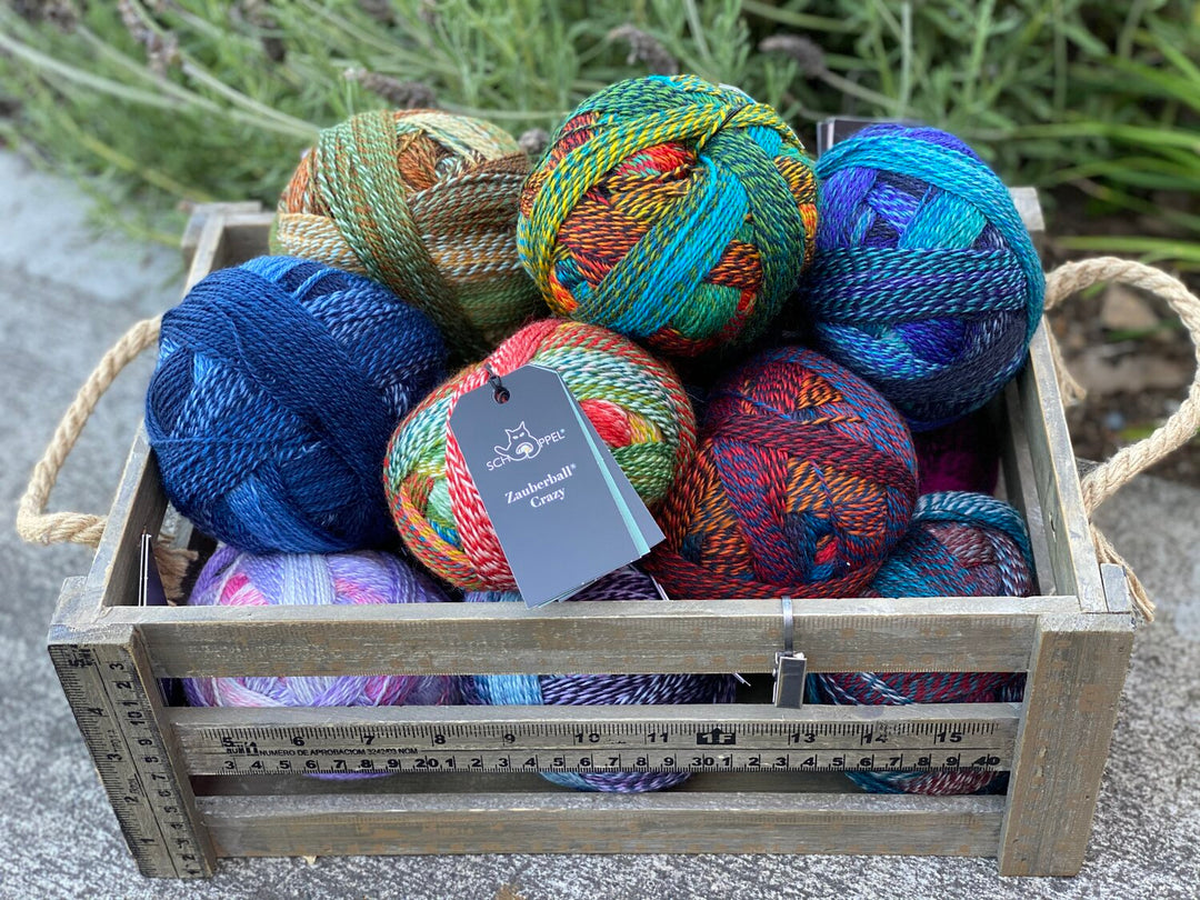 A collection of colorful self-striping yarn balls displayed in a wooden crate with a label showing the brand and yarn type.
