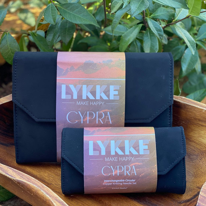Two black Lykke Cypra needle sets placed inside a wooden bowl with a plant in the background, showcasing the brand name and the product range.