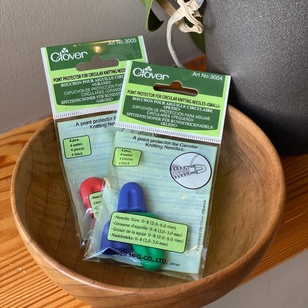 Packaging of Clover point protectors for circular needles, with one package showing small size protectors and another showing large size protectors, placed in a wooden bowl.