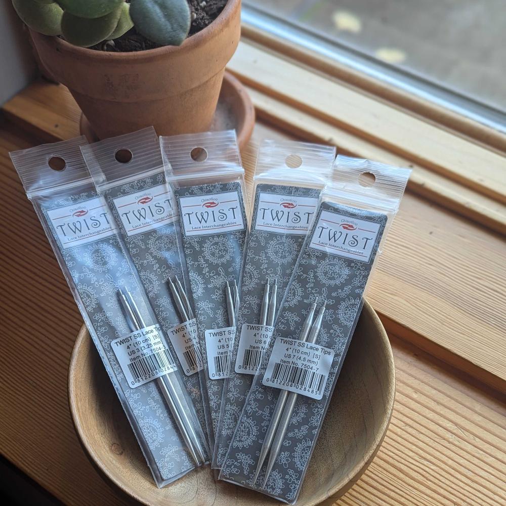 Packaged lace tips for knitting needles placed on a wooden surface with a window in the background.
