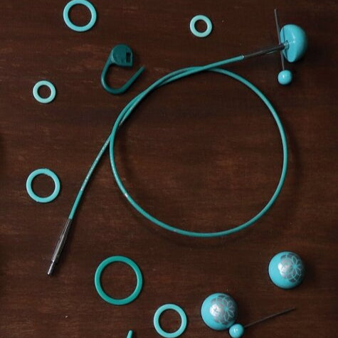 Interchangeable knitting needle cord with nylon coating, metal markings, wooden end caps, and various connectors laid out on a wooden surface.
