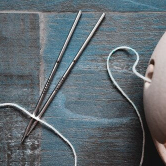 A pair of stainless steel knitting needles with lace tips, partially inserted into a ball of yarn, with a wooden surface in the background.