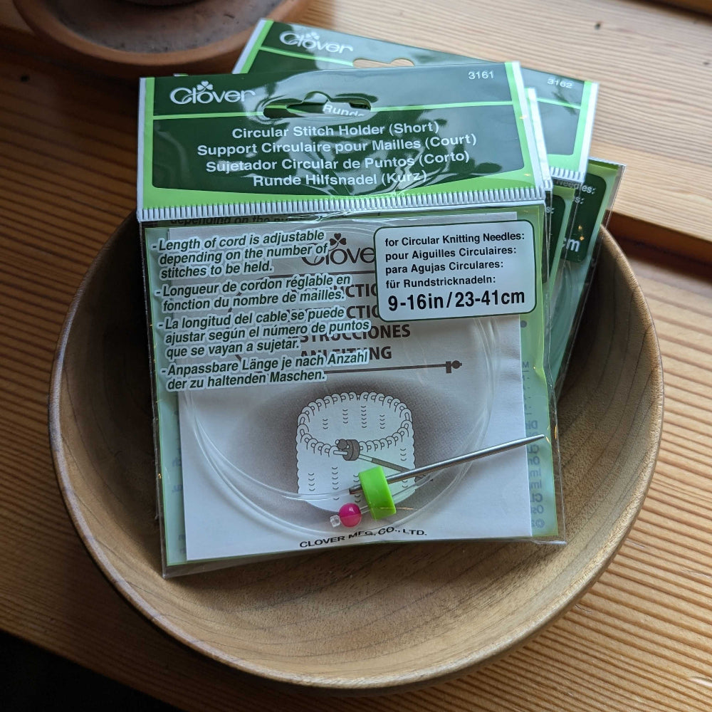 Packaging of Clover circular stitch holders displayed in a wooden bowl, with one holder partially unfolded showing the metal prongs and measurement markings.