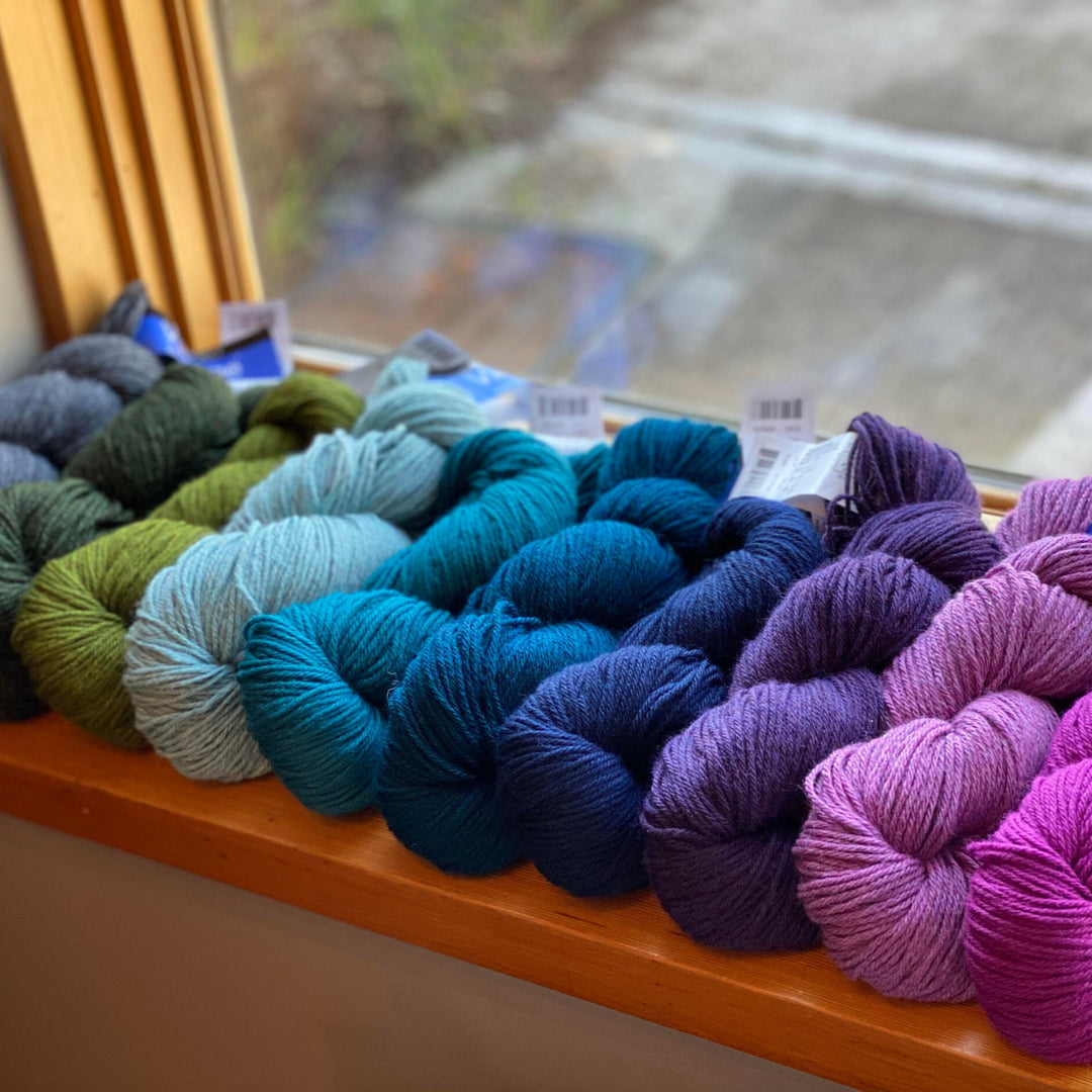Multiple skeins of Vintage yarn by Berroco arranged in a row on a windowsill, showcasing a range of colors including green, blue, purple, and pink.