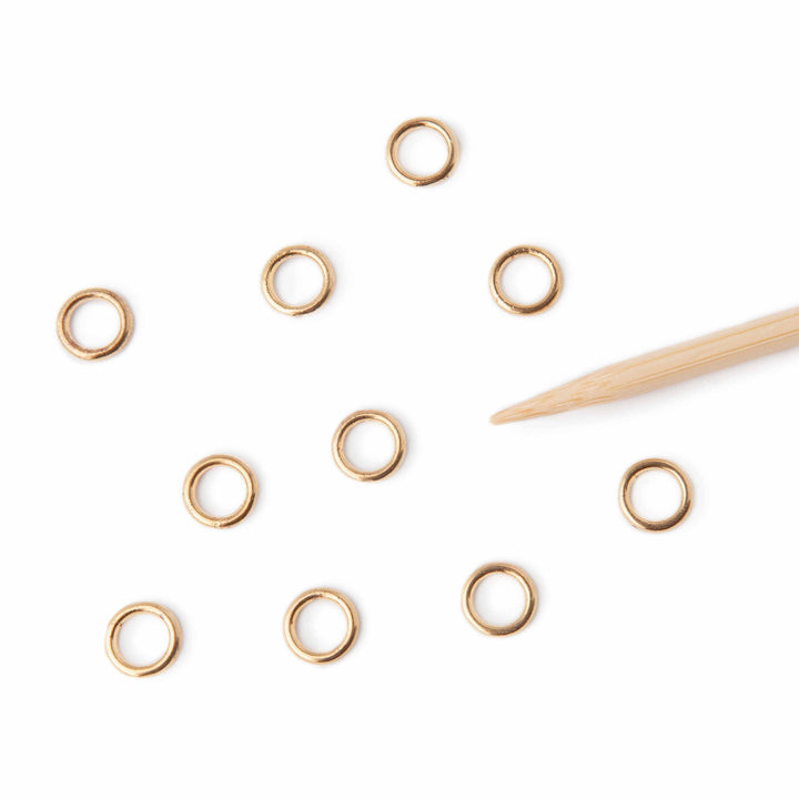 Gold stitch markers scattered on a white surface with a wooden tool.