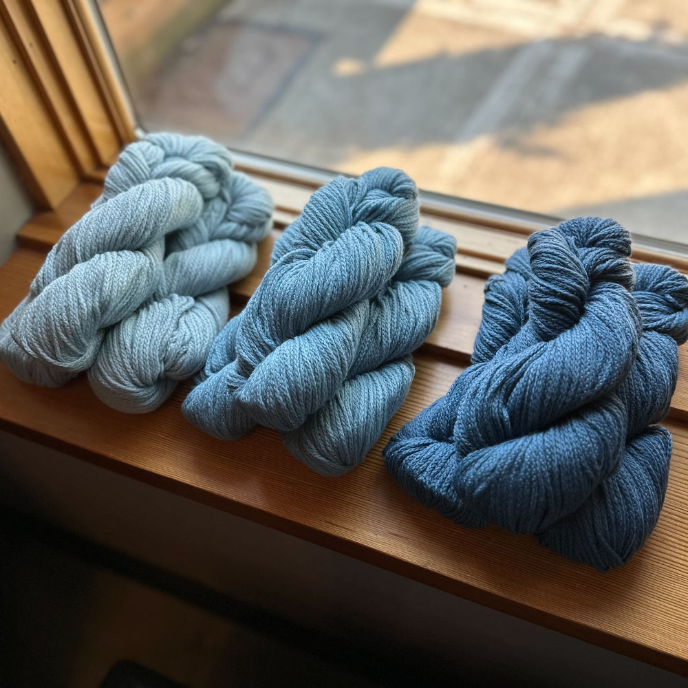 Three stacks of blue Far yarn from Woolfolk on a wooden surface near a window.