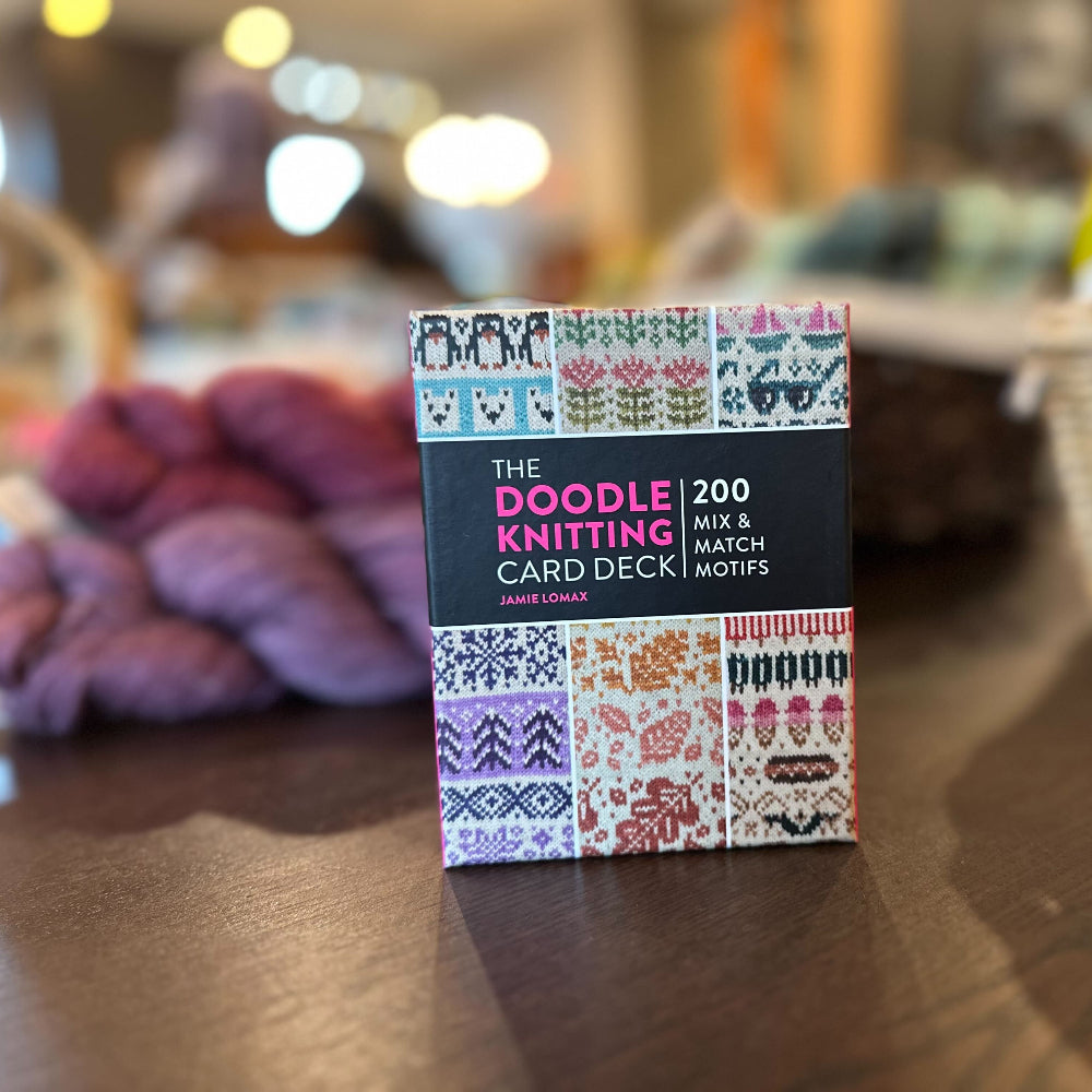 Doodle Knitting Card Deck packaging on a wooden surface with yarn and knitting needles in the background