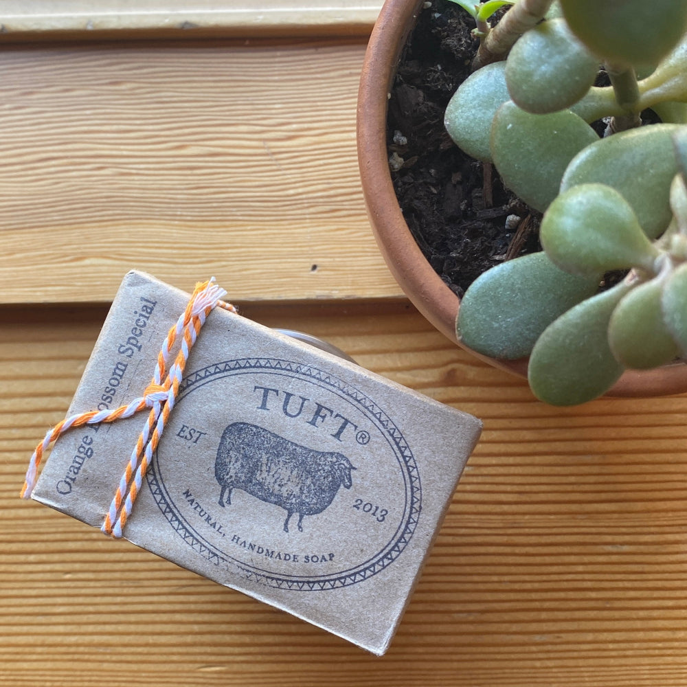 Bar of soap with a 'TUFT' label on a wooden surface next to a potted plant