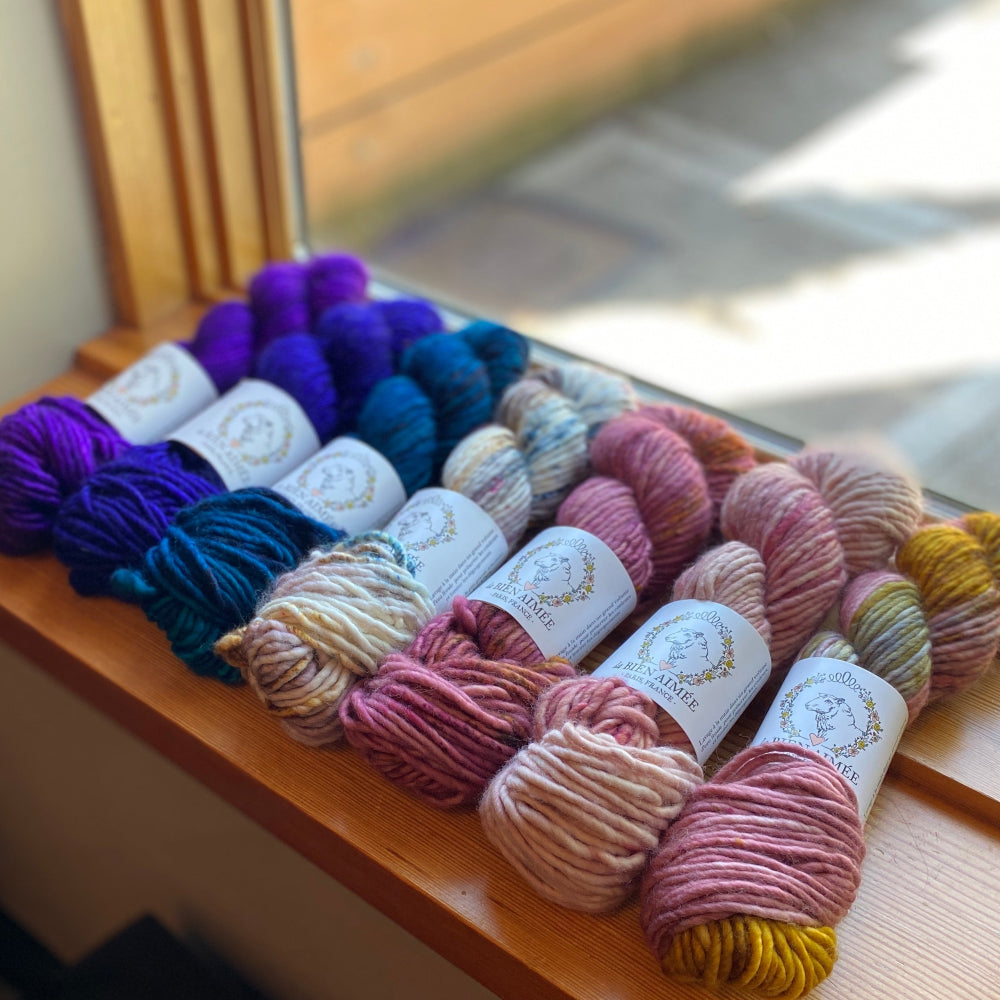 Assorted skeins of Big Merino yarn from La Bien Aimee on a wooden surface with a window in the background
