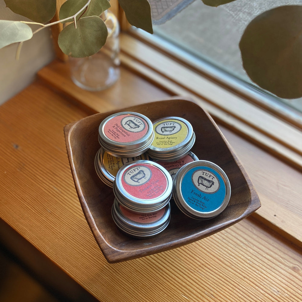 Small containers of tuft hand balm with colorful lids on a wooden tray by a window