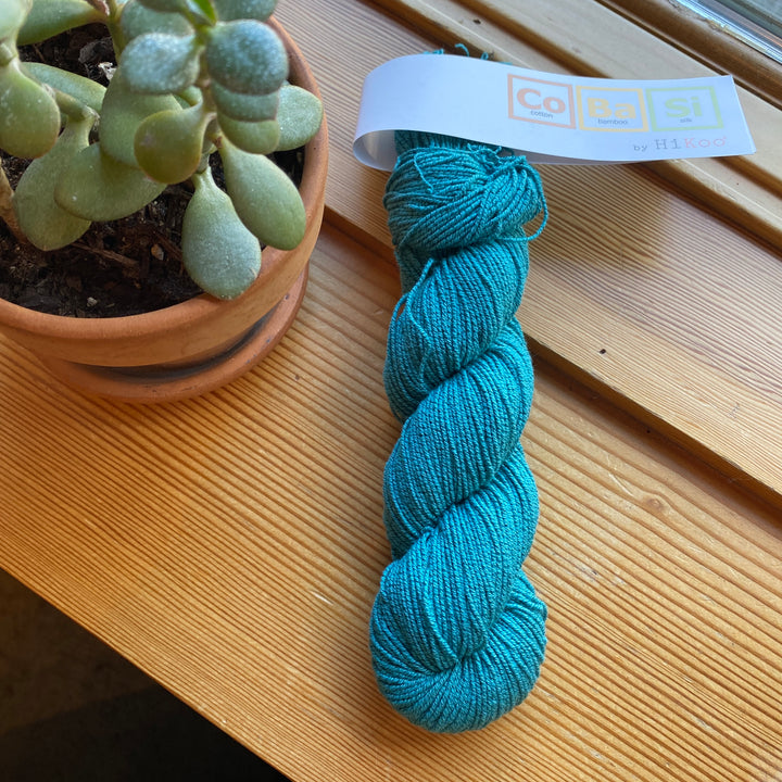 Turquoise skein of yarn with a label on a wooden surface next to a potted plant.