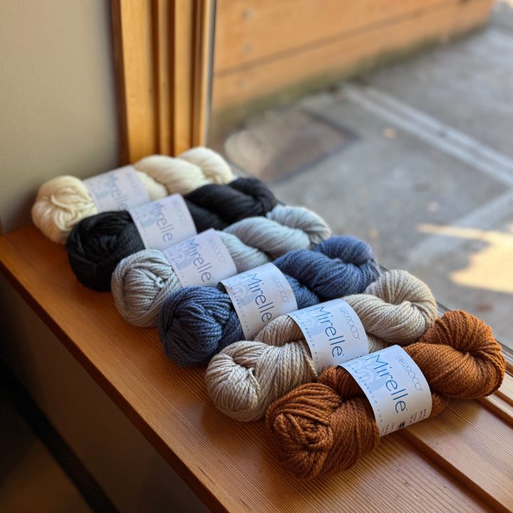Six skeins of yarn in various colors of Mirelle by Berroco on a wooden surface with a window in the background.