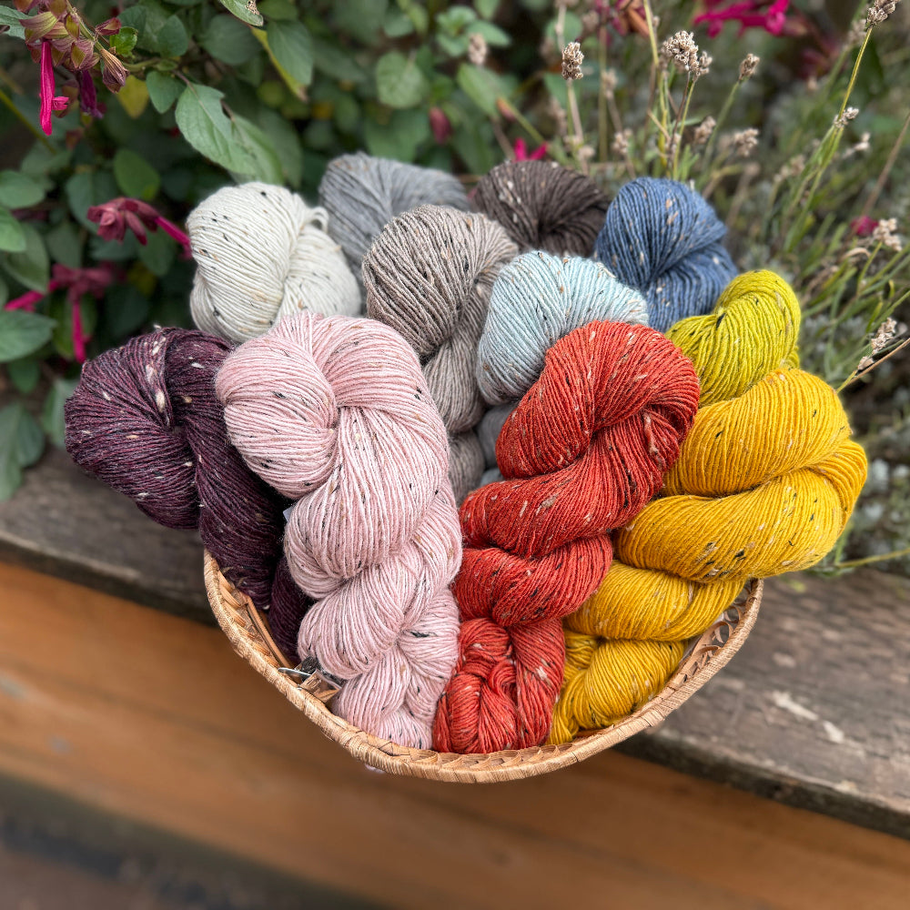 A woven basket filled with multiple skeins of yarn in assorted colors, including purple, blush pink, gray, blue, yellow, and rust red, set outdoors on a wooden surface with green foliage in the background.