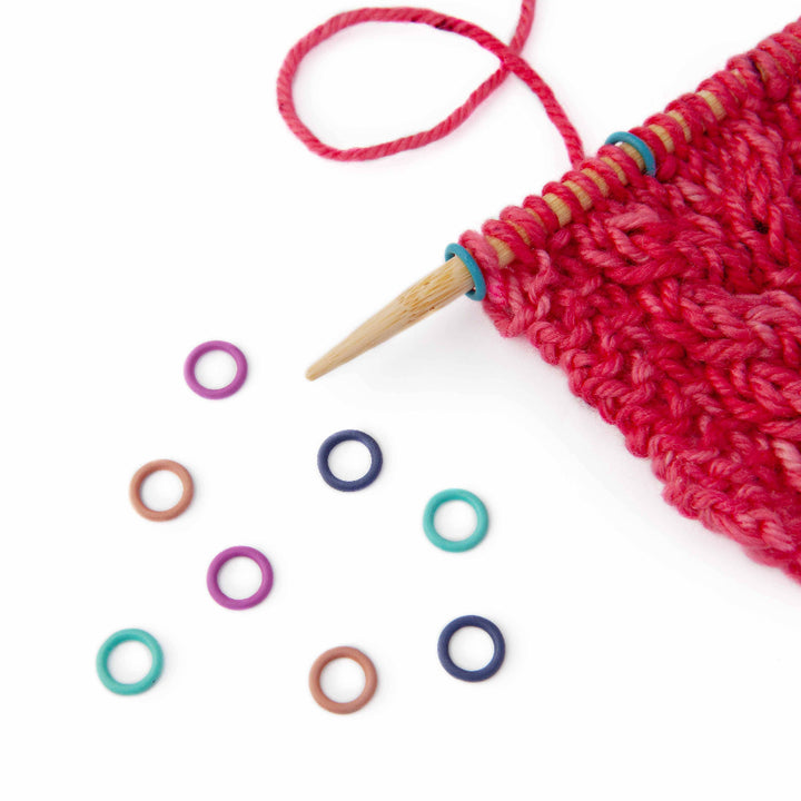Knitting project with red yarn and wooden needles on a white background, surrounded by colorful knitting markers.