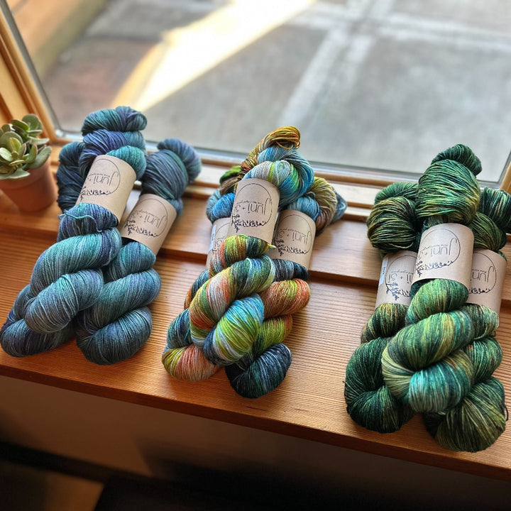 Three skeins of multicolored Juni and Si yarn on a wooden surface near a window.